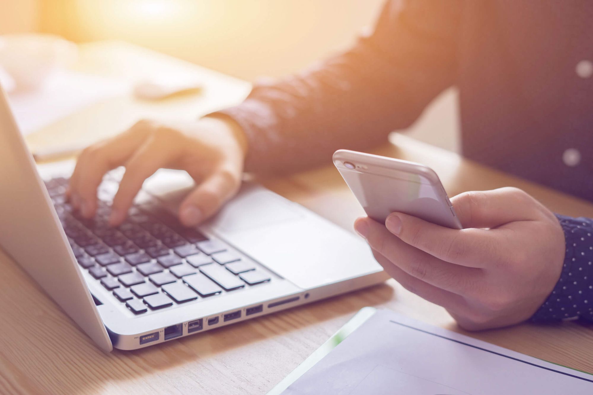 Person beim Multitasking mit Laptop und Smartphone in einem gut beleuchteten Büro.