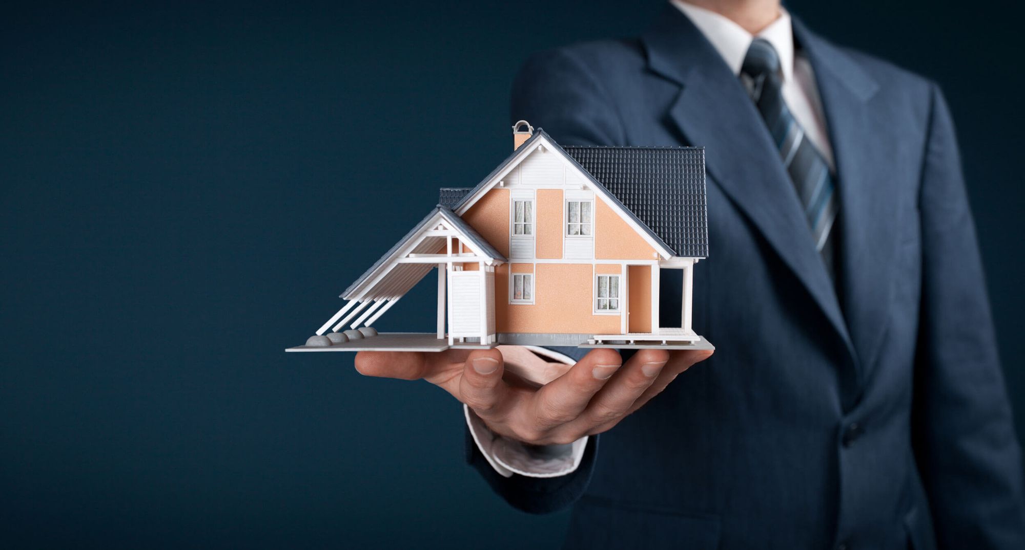 Person im Anzug, die ein Modell eines Hauses auf ihrer Handfläche vor einem blauen Hintergrund hält.
