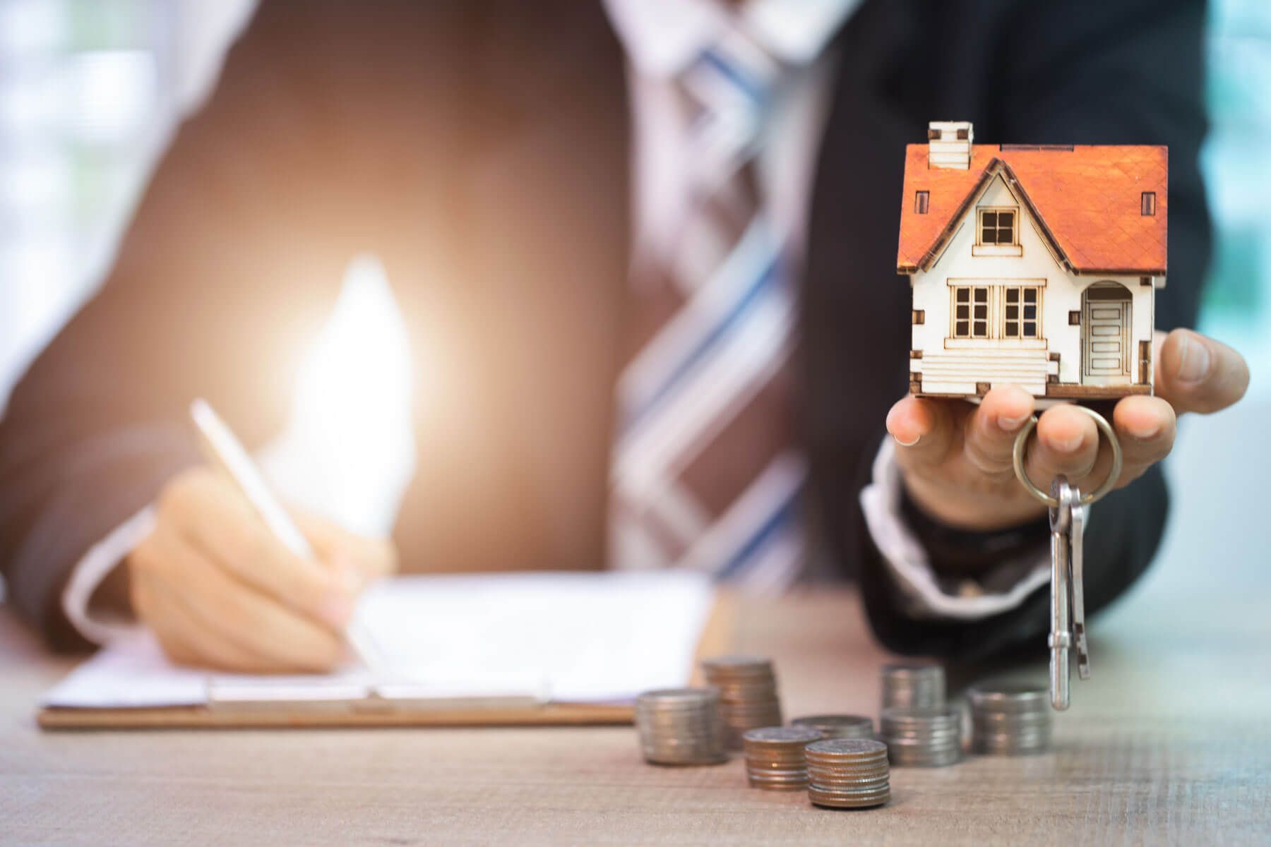 Person mit Hausmodell und Schlüsseln, mit gestapelten Münzen und Papierkram auf dem Schreibtisch.